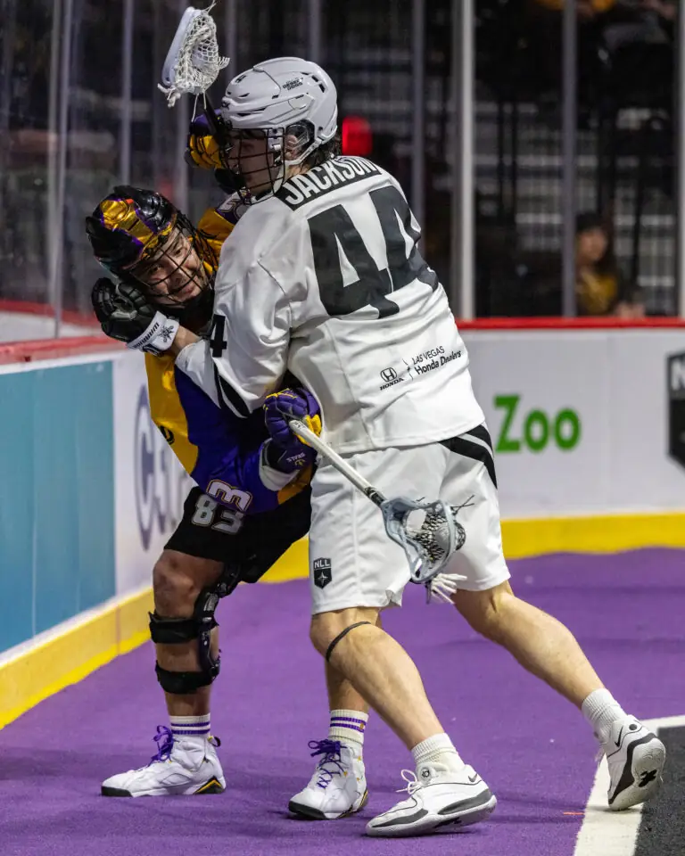 A player in a white lacrosse uniform is checking an opponent in purple against the boards during a game.