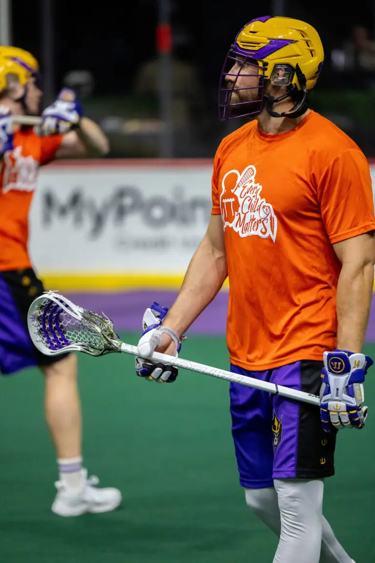 A lacrosse player in an orange "Every Child Matters" shirt holds a stick, wearing a purple and yellow helmet and gloves.