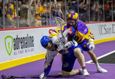 Two lacrosse players grapple on the field, one wearing a blue helmet and white jersey, while the other in a purple and yellow jersey applies pressure from behind. Fans cheer in the background.