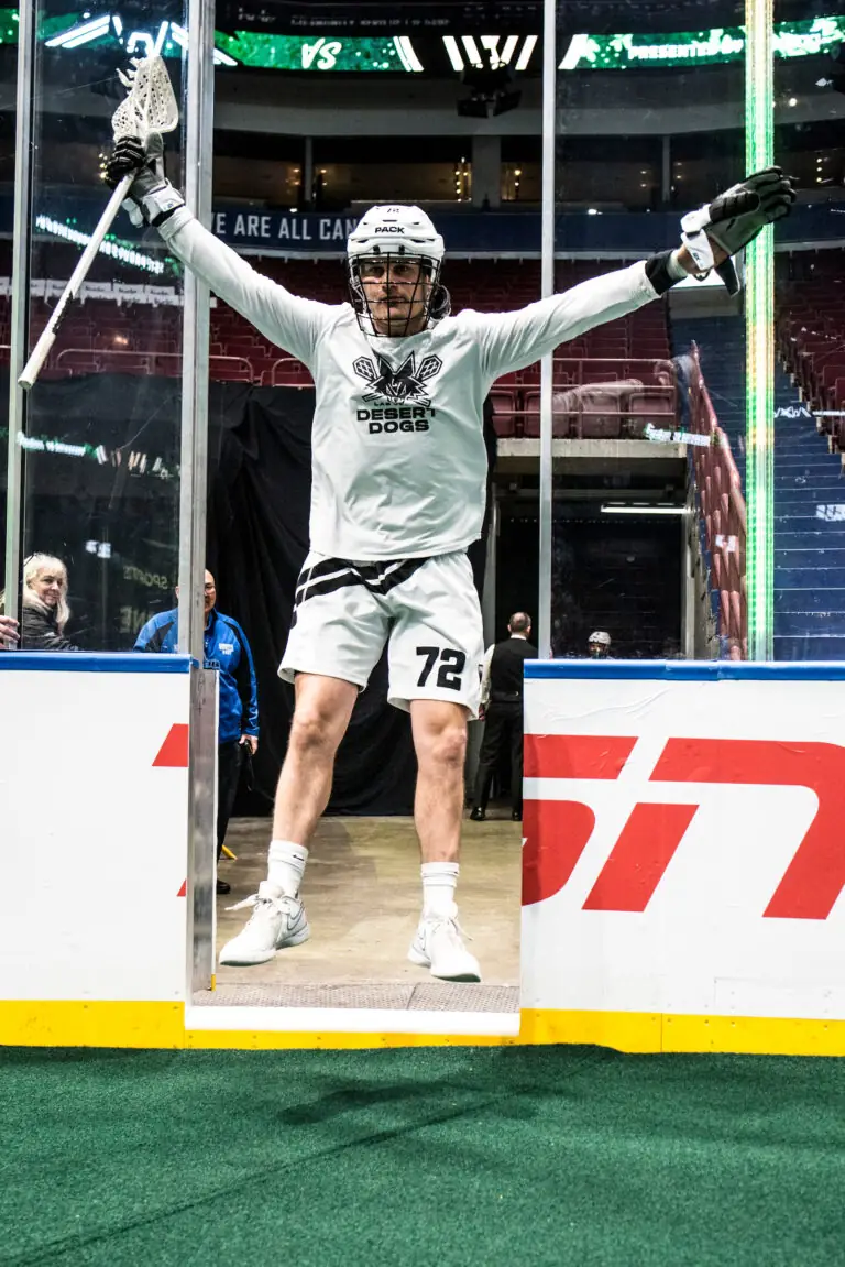 Lacrosse player in a white uniform stands at the entrance to the field, arms raised, ready to enter the game.