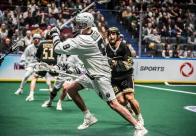 Lacrosse players compete on a green turf field, with one player in white gearing up for a shot while another in black defends.