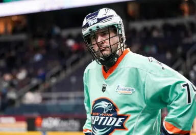 Player in a turquoise lacrosse jersey with "New York Riptide" logo, wearing a helmet and looking serious on the sidelines.