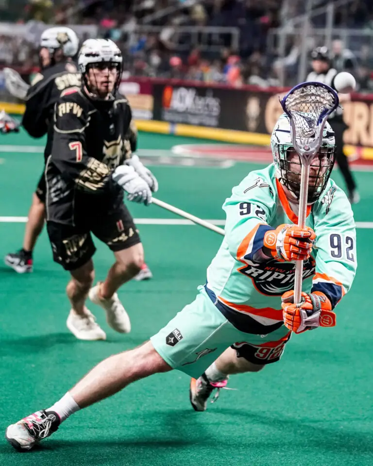 A lacrosse player in a turquoise jersey lunges to catch the ball during a National Lacrosse League game, while another player in a black jersey observes.