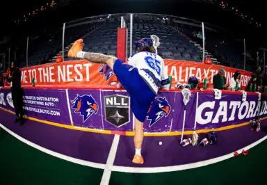 A lacrosse player stretches while wearing a team uniform, with a game bench and spectators in the background.