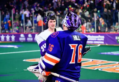 Two lacrosse goalies in conversation on the field, one in a white jersey and the other in a blue jersey marked "HILL." Fans are visible in the background.