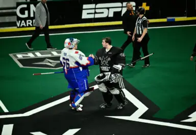 Lacrosse goalies shake hands on the field after a game, with a referee and team members in the background.