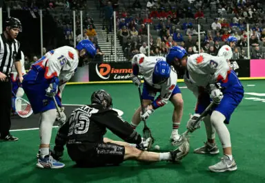 Lacrosse players compete for the ball on the field, with one player sitting on the ground and three others leaning over him. A referee watches in the background.