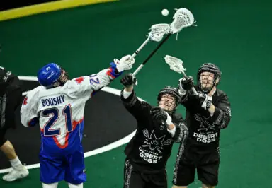 Lacrosse players battle for the ball, with one player in a white jersey reaching high while two players in black jerseys jump to intercept.