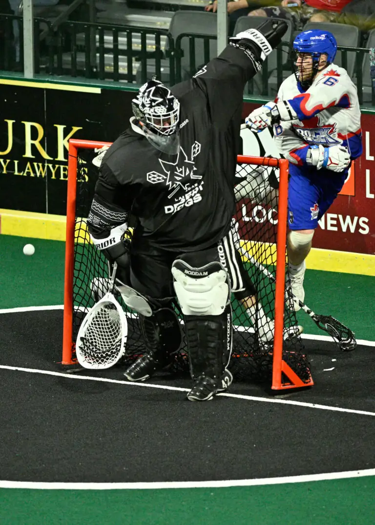 A lacrosse goalie in a black uniform celebrates a save as a player in a blue and white uniform approaches.