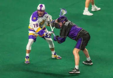 Two lacrosse players compete on a green indoor field, one in a white and purple uniform, the other in a black and purple uniform, both engaged in a stick battle.
