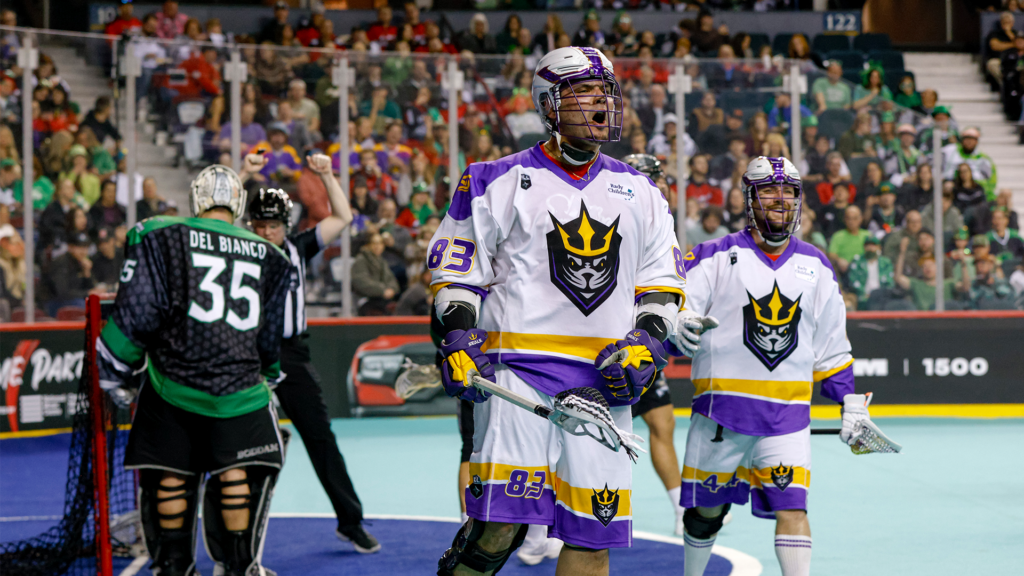 Players in colorful lacrosse uniforms celebrating during a game. The scene includes a goalie and a referee signaling a play.