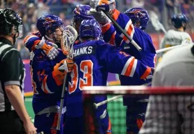 Lacrosse players celebrate a goal, wearing purple and orange uniforms, with a referee in the foreground.