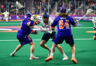 Two players in blue jerseys collide with a white-jerseyed player in a National Lacrosse League game on a green field.