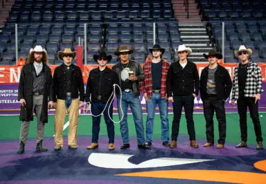 A group of eight men dressed in cowboy attire, including hats and denim, pose on a lacrosse field.