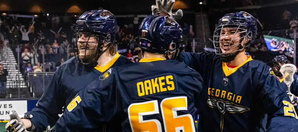 Three Georgia Swarm lacrosse players celebrate together on the field, smiling and wearing navy jerseys with yellow accents.