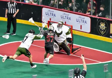Lacrosse players compete as one shoots towards the goal while the goalie prepares to defend.