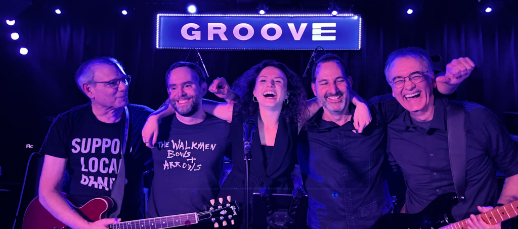 Five musicians smiling and posing together on stage at a venue called Groove, with guitars in hand and a vibrant backdrop.