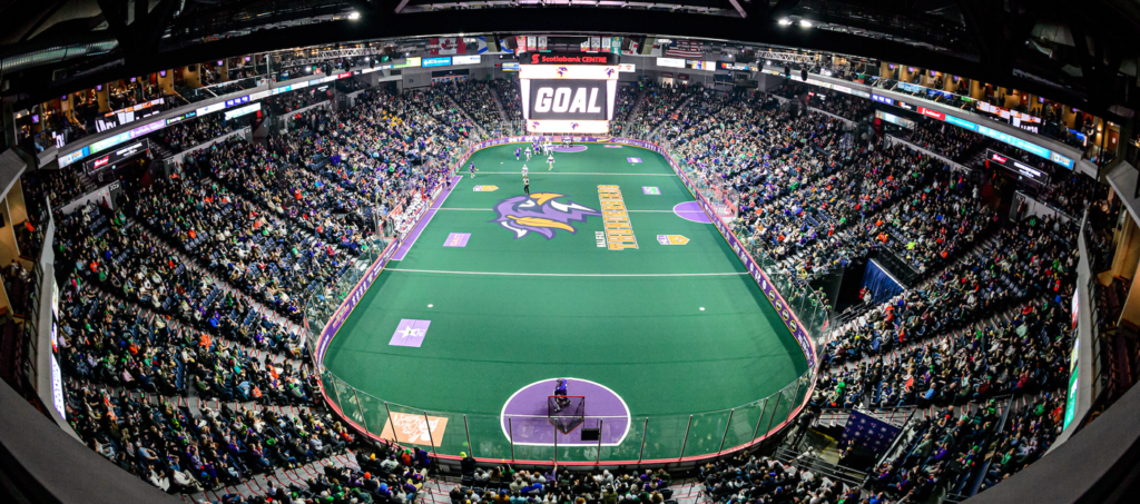 A packed arena with fans watching a National Lacrosse League game; a large scoreboard displays "GOAL."