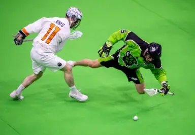 Two lacrosse players in action on a green field, one in a white jersey and the other in a bright green jersey, chasing a ball.