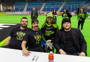 Four individuals pose for a photo at a National Lacrosse League event, with two players in team gear, a young fan wearing a jersey, and another person in a black suit. A lacrosse stick is held by the fan.