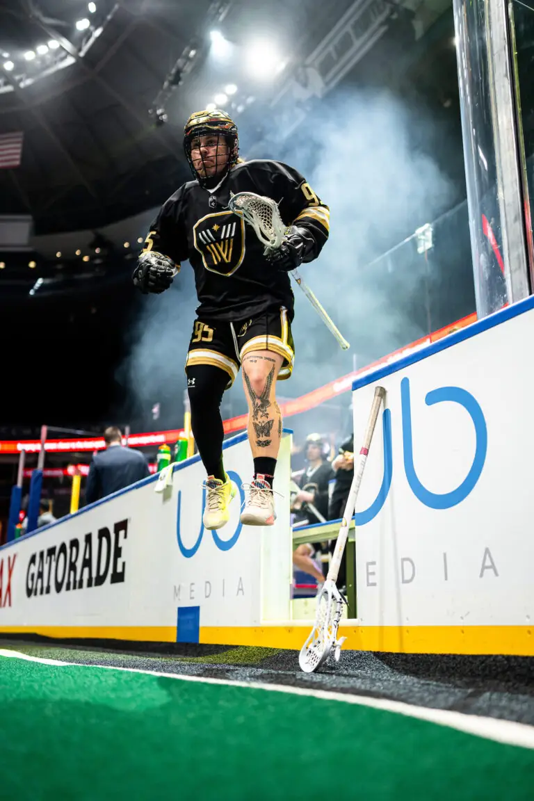 A lacrosse player in a black and gold uniform jumps onto the field, holding a stick, with smoke and bright lights in the background.