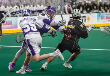 Two lacrosse players in white uniforms defend against a player in a black uniform during a game on a green field.