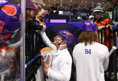 A lacrosse player in a helmet interacts with fans at a game while another player stands nearby.