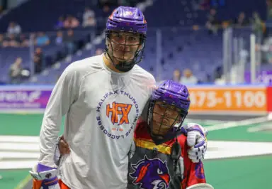 A lacrosse player in a Halifax Thunderbirds jersey poses with a young fan wearing a colorful lacrosse uniform, both smiling on the field.