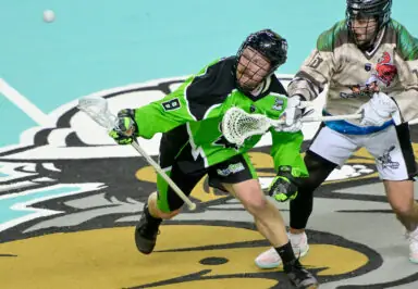 A player in a green lacrosse jersey attempts to scoop up a ball on the floor while being closely guarded by an opponent in a beige jersey.