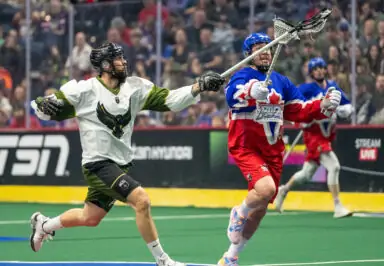 Two lacrosse players competing for the ball during a match, one in a white jersey and the other in a blue and red jersey.