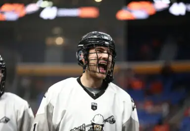 A lacrosse player in a white jersey and helmet smiles and shouts, with teammates in the background.
