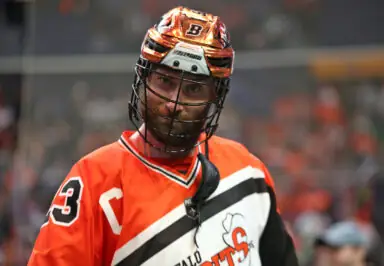 Lacrosse player in an orange jersey with a helmet, looking directly at the camera.