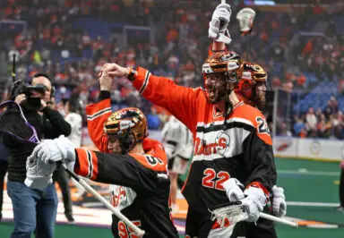 Lacrosse players celebrate a victory on the field, with fans cheering in the background.