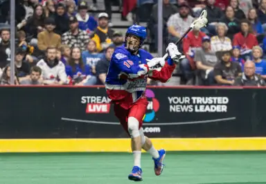 A young lacrosse player in a blue and red uniform prepares to pass the ball, with a crowd in the background.