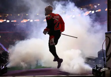 A lacrosse player in an orange and black jersey jumps through a cloud of smoke during player introductions.