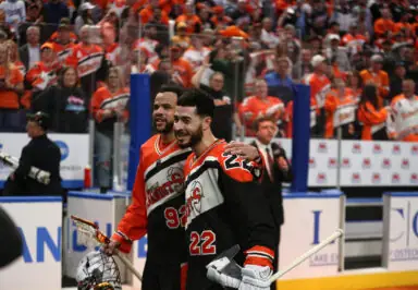 Two lacrosse players in team jerseys celebrate with a crowd of fans in the background, all wearing orange.
