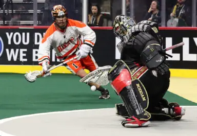 A lacrosse player in an orange and white uniform shoots the ball towards a goalie in black gear during a National Lacrosse League game.