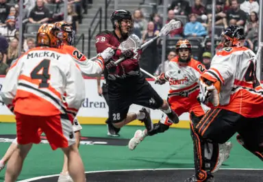 A player in black jumps to shoot the ball while being defended by players in orange and white uniforms during a lacrosse game.