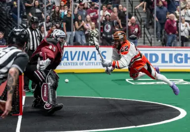 A lacrosse player in an orange and white uniform dives to make a play while a goalie in maroon gear prepares to defend the goal, with fans in the background.