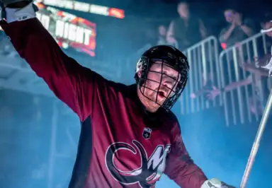 A lacrosse player in a maroon jersey and helmet gestures enthusiastically in a smoky arena, with fans applauding in the background.