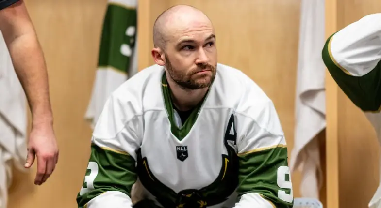 A lacrosse player in a white jersey sits in a locker room, looking thoughtful.