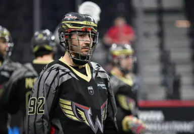 Player in black and gold lacrosse uniform, wearing a helmet and standing on the field, with teammates in the background.