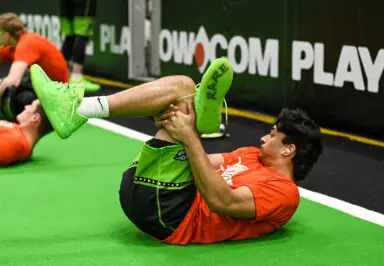 A young athlete stretches on a green mat, holding one leg while lying on his back, with teammates visible in the background.