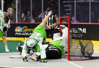 A player in green dives into the goal, colliding with the goalie in a National Lacrosse League game, while another player looks on.