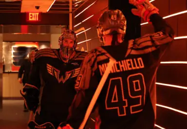 Two lacrosse players in black jerseys pose in a brightly lit hallway, one waving while the other looks on.