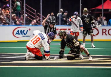 Two lacrosse players face off at center field during a game, with a referee overseeing the match.