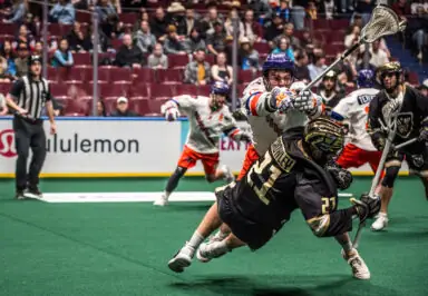 A lacrosse player in a black uniform is tackled by an opposing player in a white uniform during a National Lacrosse League game.