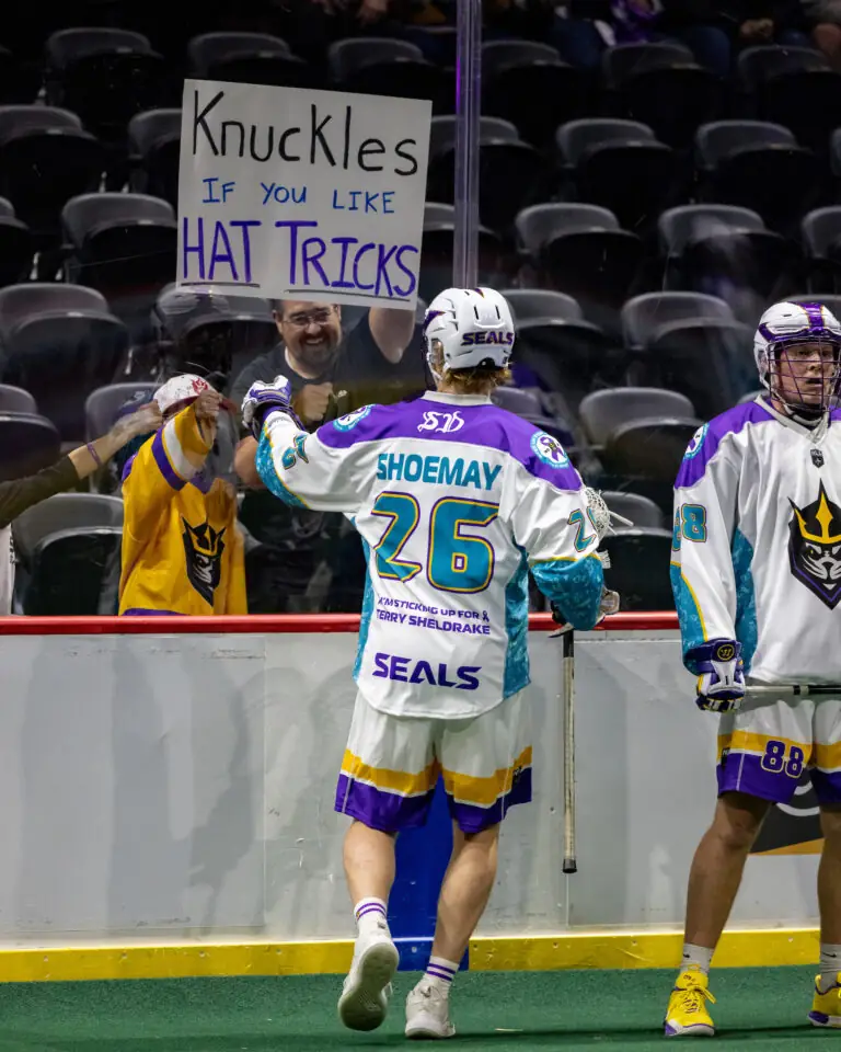 A lacrosse player in a blue and white jersey, labeled "SEALS," interacts with a fan holding a sign that says, "Knuckles if you like hat tricks."
