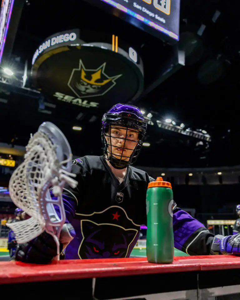 A lacrosse player in a black uniform with a panther logo prepares on the sidelines, holding a stick and a water bottle.