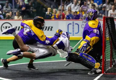 A player in a yellow and purple jersey dives towards the goal, colliding with a defender in white, while a goalie in purple prepares to block the shot.